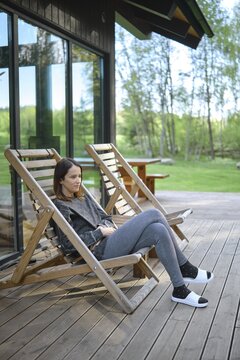 A woman sits comfortably in a wooden deck chair, enjoying the tranquil atmosphere. The lush green landscape and bright sunlight create a serene environment, perfect for relaxation