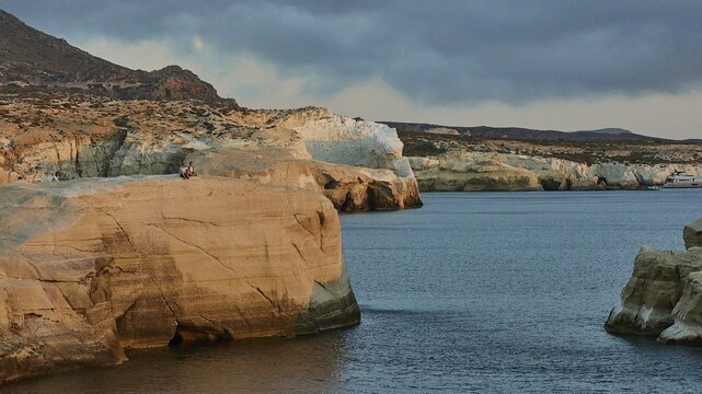 Morning light, tuff rock, tuff rocks, 2 people sitting on rocks, calm dark blue sea, grey-blue clouds, Sarakiniko Beach, Milos Island, Cyclades, Greece