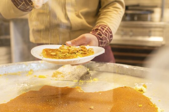 Close up kunafa dessert being served in paper plate with hot sugar syrup poured from metal bowl street sweets shop food preparation close up. Popular middle east and mediterranean dish