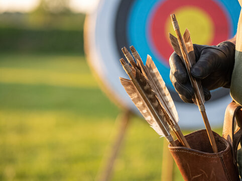 hand in a black leather glove pulling feathered arrows out of a leather quiver against a colorful archery target outdoors