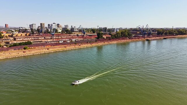 A small boat moves along the river with the city skyline and industrial buildings in the background. It is a clear day with no clouds in the sky.
