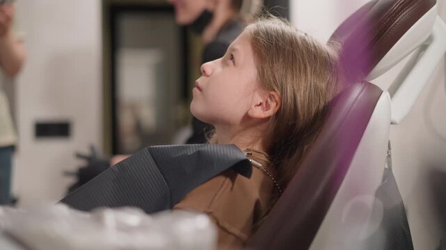 Close up of young girl sitting in dental chair during orthodontic consultation. Dental bib covers her chest while another girl stands nearby indicating professional setting with soft