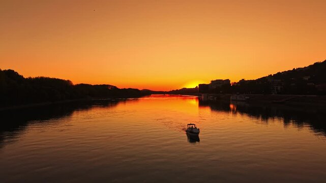 A boat travels across the water as the sun sets in the background. The sky is orange and reflects off the river surface. Trees and buildings line the riverbank.