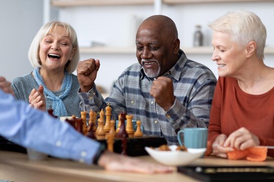 Smiling Senior Group Of Three Members, Mixed Gender And Ethnicity, Playing Chess With Excitement At Cozy Home Setting. Engaging Activity Bringing Joy And Connection Among Friends.