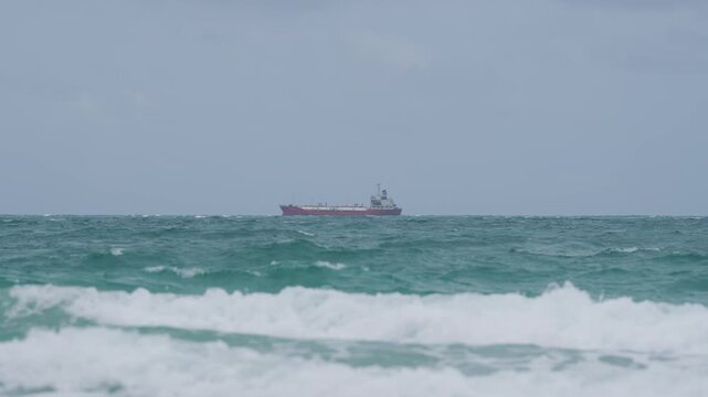 Rough ocean wave cargo ship on storm sea near horizon, turquoise water with white foam surf, distant freighter tanker crossing moody overcast sky seascape, maritime transport shipping travel scene