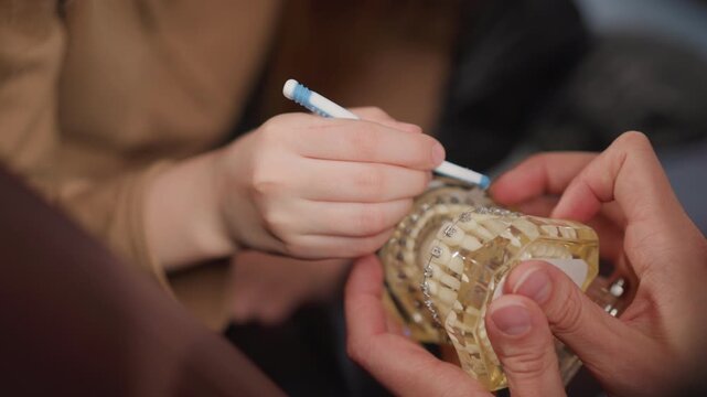 Close up of young person examining dental braces on orthodontic model while another hand holds clear plastic mold. interaction focuses on pointing out specific teeth with visible metal
