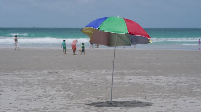 Beach ocean umbrella sand summer vacation seaside shoreline, colorful parasol on empty coast, distant family near waves, calm wave, blue sky horizon, travel relaxation background copy space