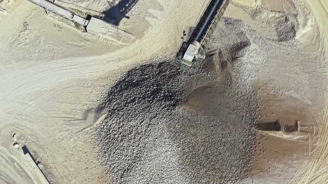 A drone captures a view of a construction site with a large pile of gravel in a sandy area. Machinery is visible, working to move materials during the day.