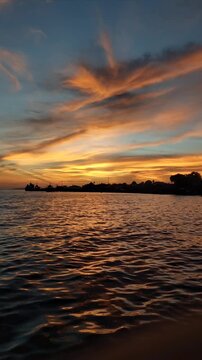 Vertical view of a dramatic sunset sky with sweeping fiery orange clouds reflecting on gentle ocean waves over a silhouetted coastal village in Biak, Papua, Indonesia.