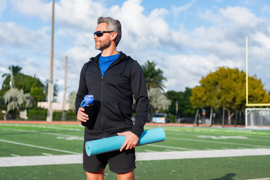 Fit your body. Yoga coach. Sport man after workout. Healthy lifestyle. Fitness and sport. Yoga mat for training. Sportsman with water bottle. Man in sportswear with fitness mat in stadium