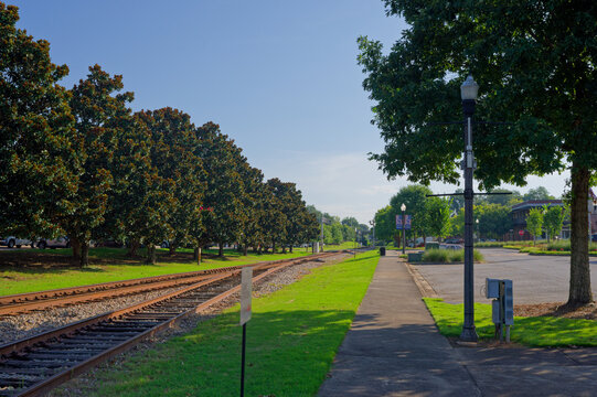 Sunlit railroad tracks run parallel to a tree-lined sidewalk in a quiet small town in Opelika, Alabama framed by mature shade trees, lampposts, green lawns and clear blue sky for a peaceful suburban s