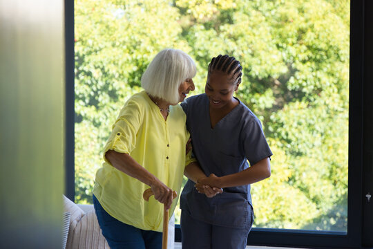Senior woman, African American caregiver in gray scrubs guiding steps with cane at home near window