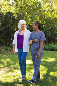 Senior and African-American caregiver in scrubs walking arm-in-arm in park with smartwatch necklace