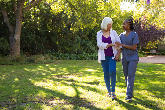 Diverse female caregiver in gray scrubs guiding senior woman across sunlit garden under shade tree