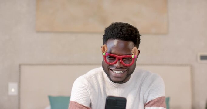 African man holding phone with novelty red glasses, laughing while watching funny clip in bedroom