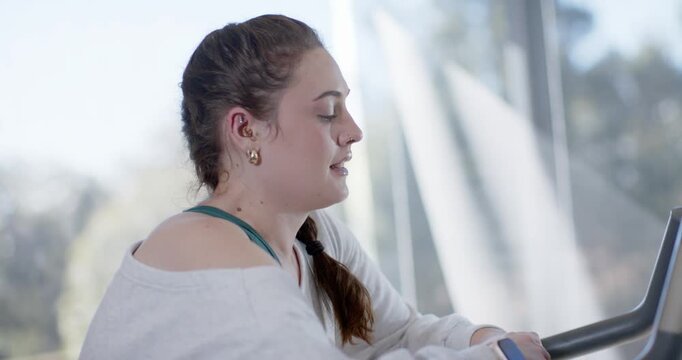 Adult female in teal tank sitting on elliptical at gym, smiling tapping smartwatch