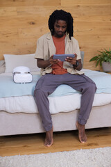 African American man sitting on bed using tablet in bedroom with VR headset, wearing beige shirt © WavebreakMediaMicro
