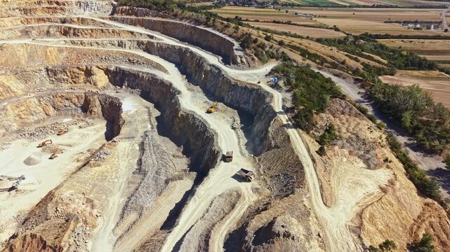 A large quarry site is shown with deep cuts in the ground. Heavy machinery is present, working on stone extraction. The location has flat lands surrounding the quarry.