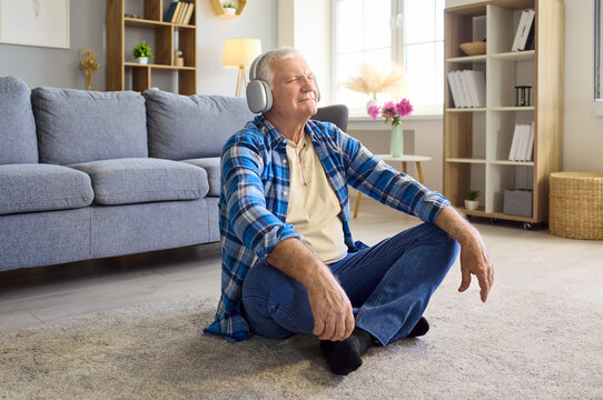 Senior man headphones meditation relaxing at home. Retiree seated on carpet, eyes closed, listening to music with headphones for wellness and relief. Calm mindful leisure lifestyle concept.