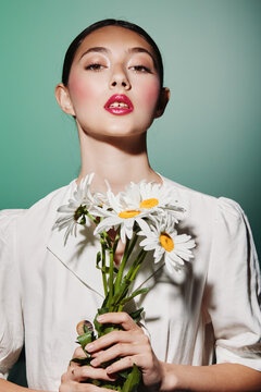 Young woman with glossy red lips and flawless skin holds a bouquet of daisies wearing a white blouse against a soft green background. Creative fashion beauty concept.