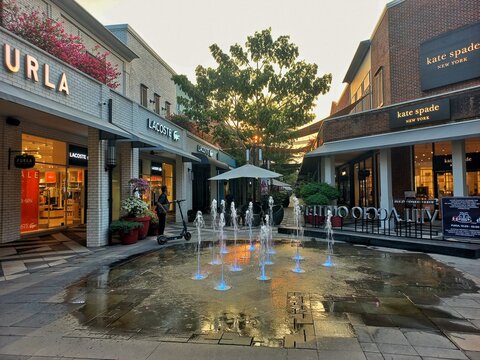 Karawang, Indonesia - August 11, 2026: Illuminated fountain and luxury retail storefronts of Furla, Lacoste, and Kate Spade at Summarecon Villaggio Outlets outdoor mall.