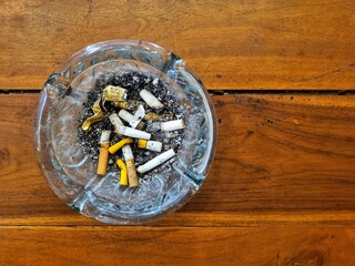 Top view of a glass ashtray filled with cigarette butts on a textured wooden background. © RJuniantoro_