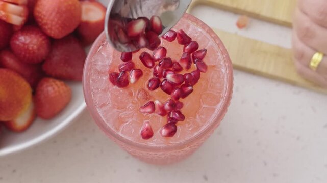 Top view of woman adding fresh pomegranate arils with a spoon to an iced pink strawberry cocktail in a textured glass