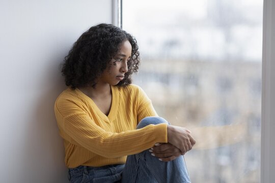 Worried Young Black Woman Sitting By Window, With Her Knees Hugged To Her Chest, Lost In Thought In A Cozy Indoor Setting, Contemplating Life And Emotions. Mental Wellness Concept.