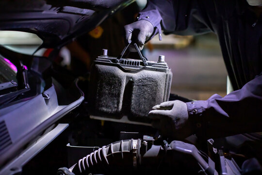 Selective focus to Professional mechanic in gloves replacing an old car battery inside an open engine compartment at an auto repair shop night service center. On-site battery replacement service.
