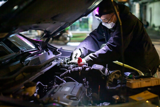 Selective focus to Professional mechanic wearing a mask and headlamp performing on-site mobile car battery replacement service at night on a broken down vehicle.