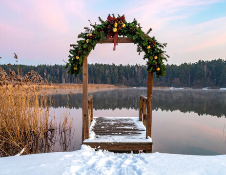 Christmas arch decorated with evergreen garland and ornaments beside snowy lakeside dock, creating peaceful winter holiday scene beneath pastel sky
