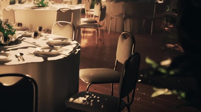 The scene shows an empty table with plates and cutlery placed on a white tablecloth. Several chairs surround the table with soft lighting casting shadows in the background.