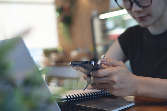 Young asian woman using smartphone while remote working on laptop with notebook on desk in cafe, modern workspace, online business lifestyle, freelance at work concept