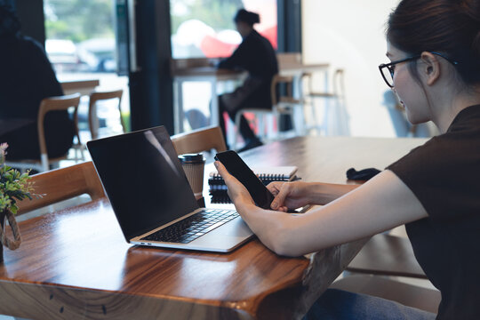 Asian woman using smartphone and laptop at cafe table, modern remote work, business casual and digital lifestyle concept.