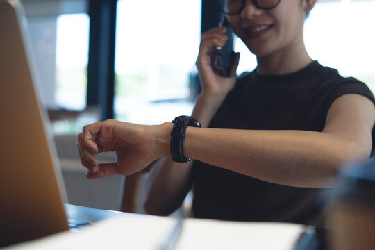 Smiling woman talking on smartphone while checking smartwatch with laptop on desk in modern workspace or home office, remote work concept.