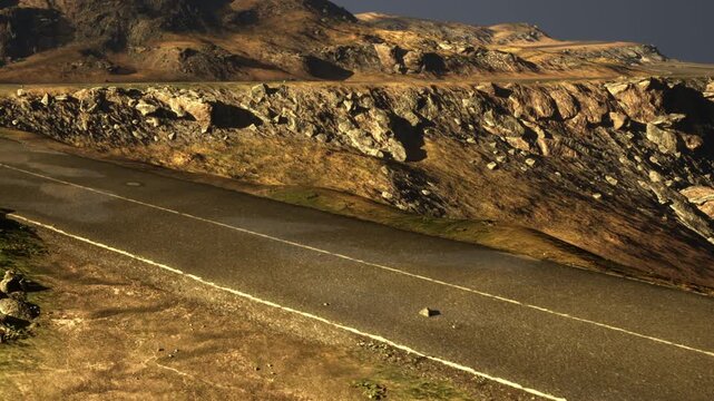 A road runs along a mountain. The ground is rocky and dry with patches of grass. Sunlight brightens the landscape. The sky is clear and blue, showing no signs of clouds.
