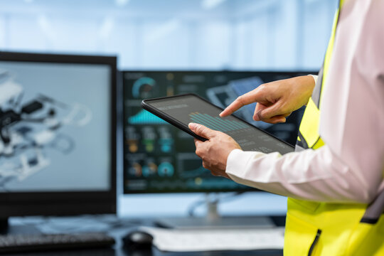 Engineer in a safety vest uses a tablet while reviewing 3d design data on computer screens in a modern industrial workplace.