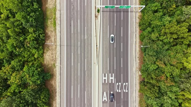 Vehicles drive on a wide, divided highway with clear lane markings. Green trees line both sides, showing a rural setting. The sun is shining on a clear day.