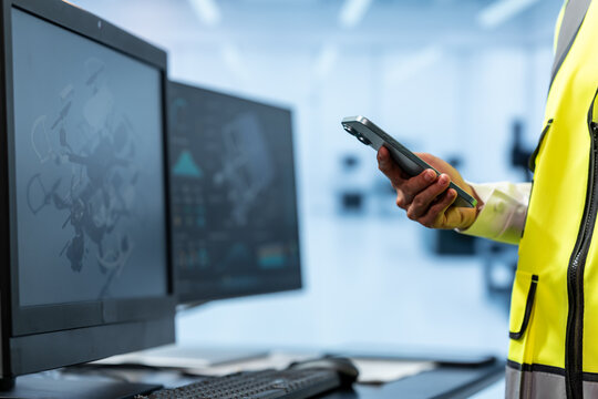 Factory worker in a high visibility vest uses a handheld scanner beside computer monitors showing industrial data in a modern control room.