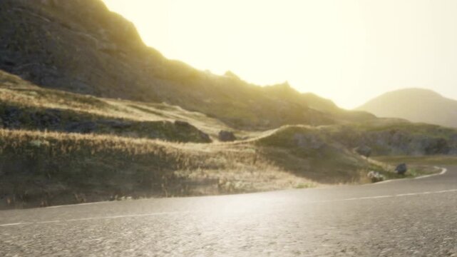 A road stretches through a grassy area while hills rise in the background. The sun is setting, casting warm light over the scene. The time is late afternoon or early evening.