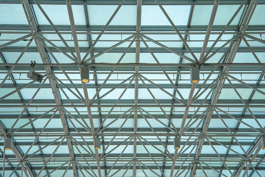 Close-up of the glass and metal canopy roof frame featuring mounted ceiling lighting fixtures and crossing pipes against the bright sky.