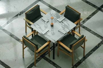 Fotobehang Horeca Top view of an empty square restaurant table with a marble top set with plates, cutlery, napkins, and four wooden chairs with green cushions.  © AncientAftertone