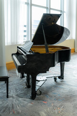 Classical black grand piano with an open lid standing on a heavy metal caster frame inside an empty room near bright glass windows. © AncientAftertone