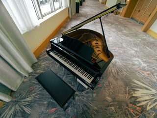 High-angle shot of a classical black grand piano with an open lid and a leather bench standing on a patterned carpet inside an empty room. © AncientAftertone