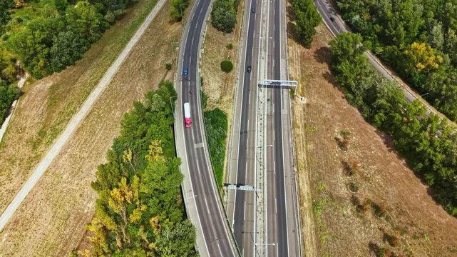 A truck drives along a curved road surrounded by green trees and open fields. The sunlight shines down on the scene, showing a mix of nature and infrastructure.