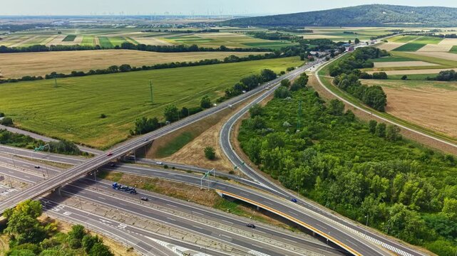 A view shows a highway intersection in a rural area. Fields stretch out on both sides and hills rise in the background. Cars move along the roads under bright daylight.