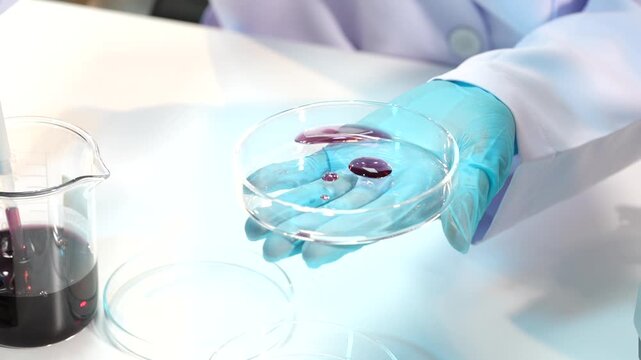 Scientist wearing blue gloves uses a pipette to place purple liquid drops in a glass petri dish beside laboratory beakers.