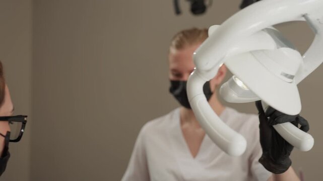 Close up of dental professional adjusting specialized oral examination light in clinical setting. Dental assistant observes procedure while patient lies prone emphasizing precision