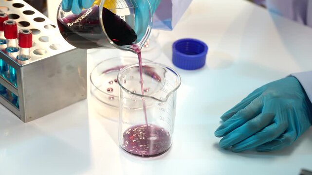 Laboratory worker in blue gloves pours purple liquid from a flask into a glass beaker beside test tubes and lab equipment.