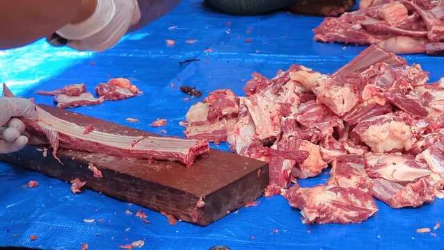 Close-up of a person wearing a gloved hand using a large cleaver to chop raw beef meat and bones on a wooden block on a blue tarp.